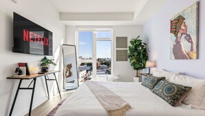 Quarto moderno com uma cama grande, televisão na parede, escrivaninha com uma planta e vista para a cidade pela janela.