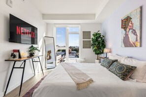 Ein modernes Schlafzimmer mit einem großen Bett, einem an der Wand montierten Fernseher, einem Schreibtisch mit Pflanze und Blick auf die Stadt durch das Fenster.
