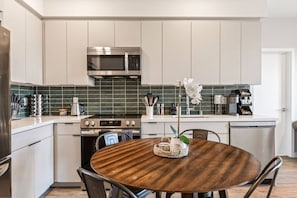 A modern kitchen with white cabinetry, a stainless steel microwave, and a round wooden dining table with chairs. - Light-Filled Modern Space in K-Town + Hot-tub & Gym (Los Angeles)
