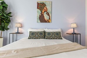 A bedroom with a bed, two nightstands, two lamps, a large poster, and a potted plant.
