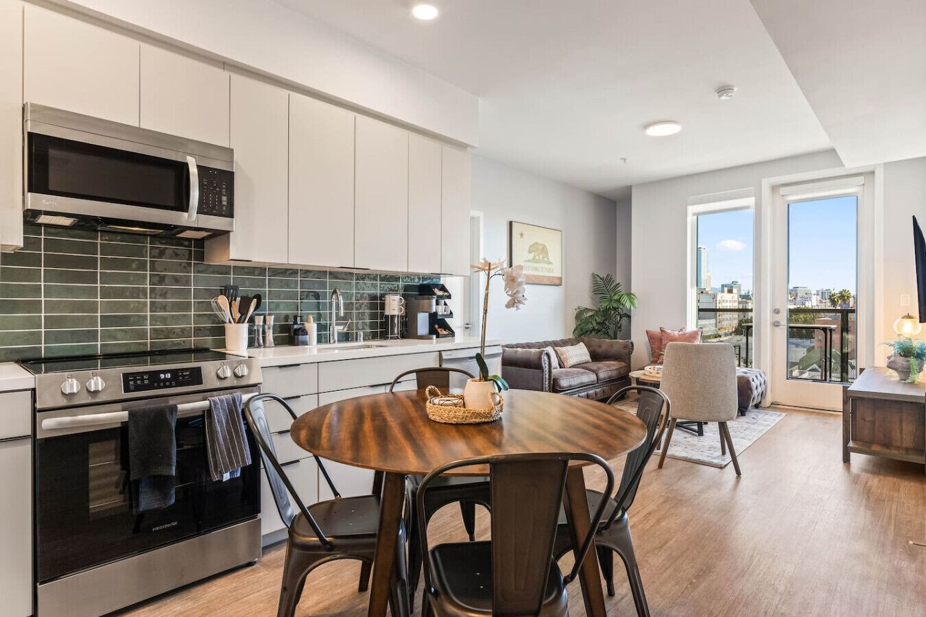 A modern kitchen with a dining area, a sofa, and a view of the city through large windows.