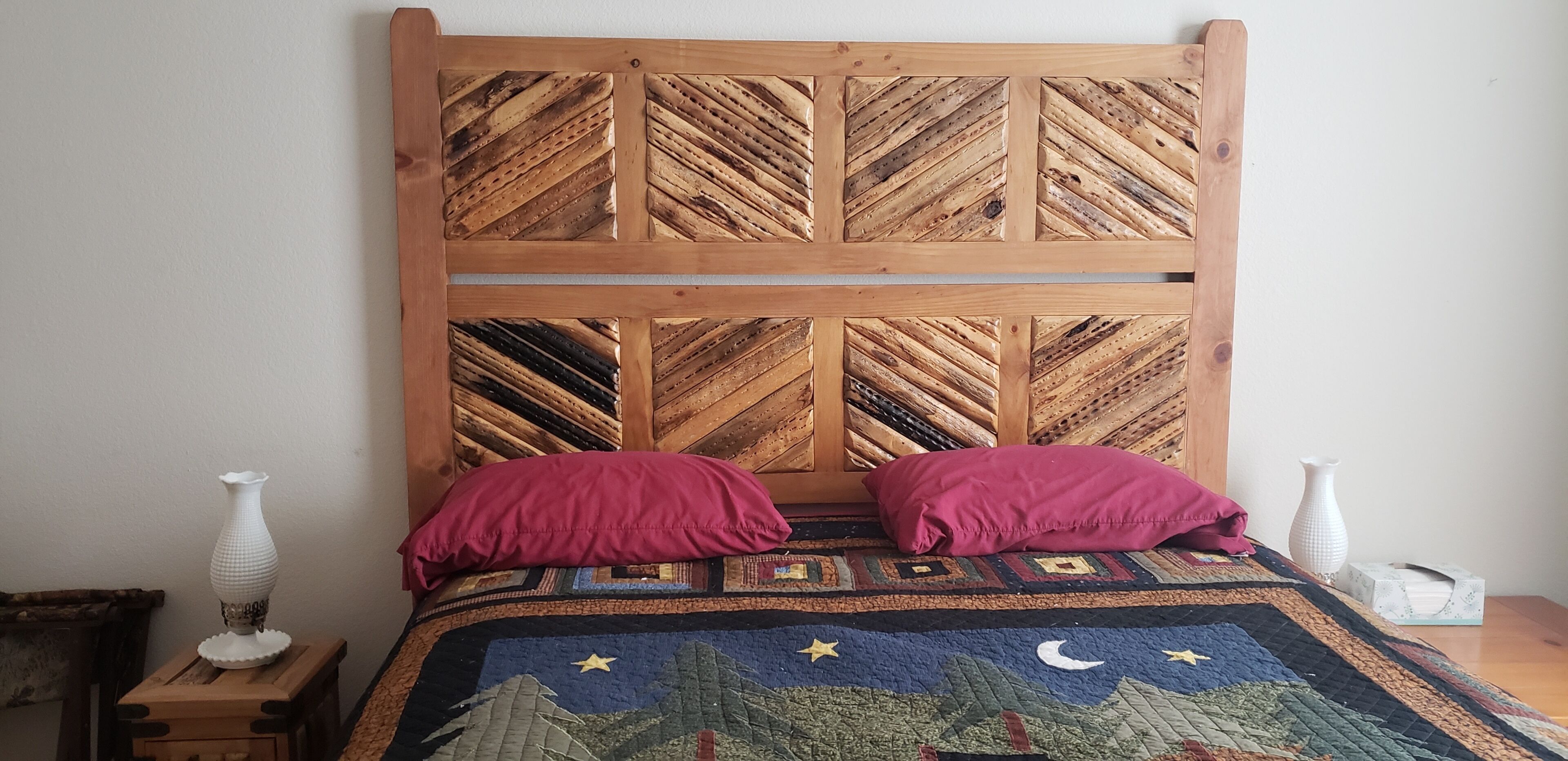 Een bed met een houten hoofdbord met een geometrisch patroon, twee kussens en een sprei met een sterrenhemel ontwerp.