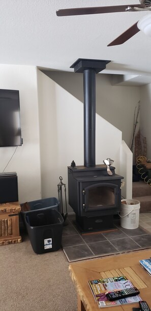 Ein Zimmer mit einem schwarzen Kaminofen, einem an der Wand befestigten Fernseher und einem Holztisch mit Zeitschriften und Fernbedienungen.