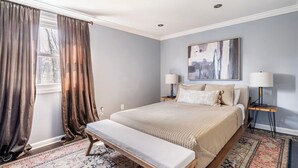 Quarto com cama grande, duas mesas de cabeceira, um banquinho, tapete e janela com cortinas.
