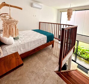 Un dormitorio con cama de madera, decoración colgante de macramé, una ventana con vistas y un balcón con plantas.