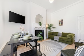 A modern living room with a dining area, a fireplace, a green sofa, and a flat-screen TV mounted on the wall. - Bayswater Two-Bed with Period Charm (London)