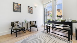 En matplats med ett bord dukat för fyra personer, två stolar, ett litet sidobord med en vas och utsikt över omgivningen genom stora fönster.