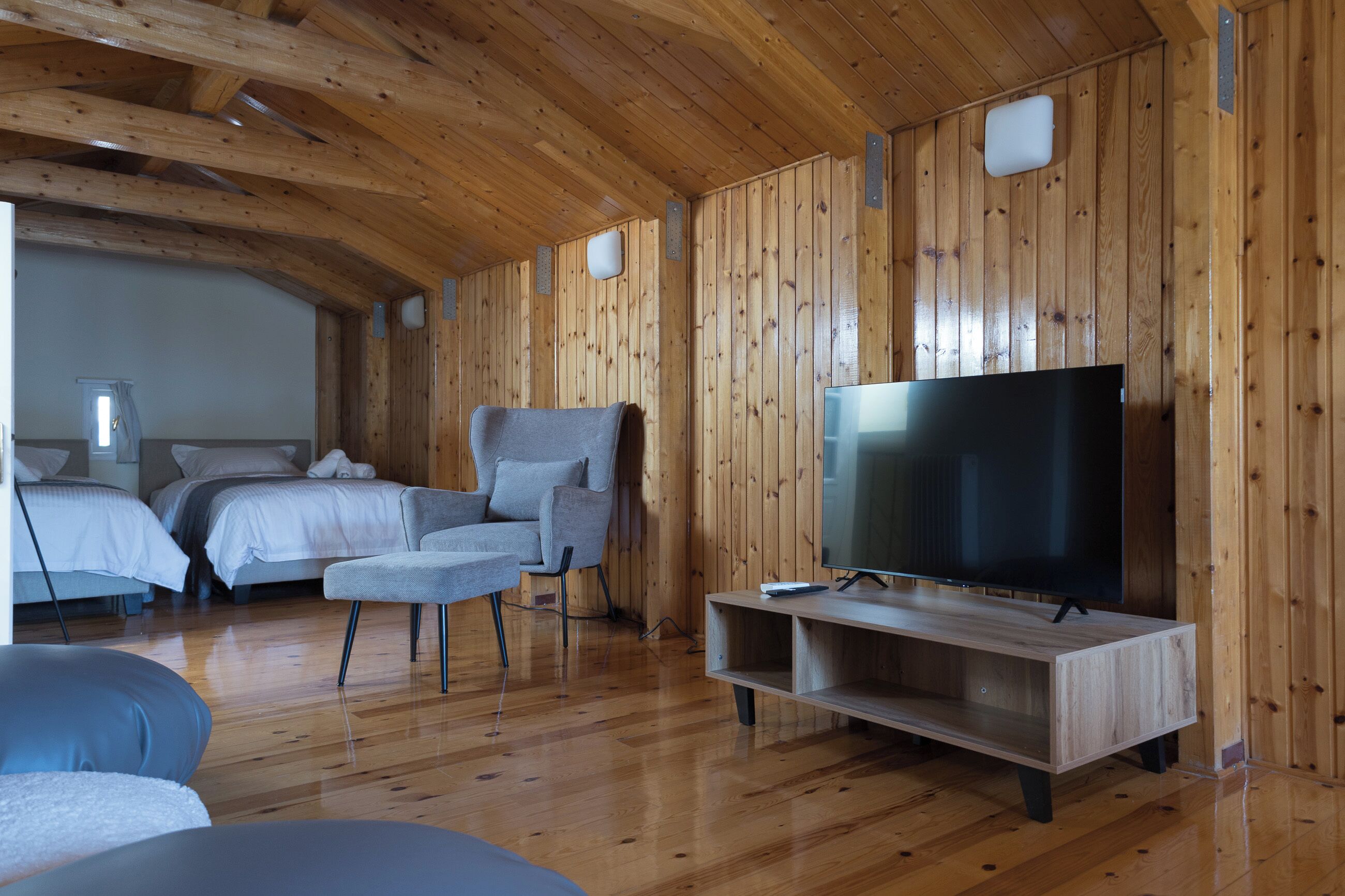 A cozy room with wooden walls, a flat-screen TV on a wooden stand, a grey sofa, and a bed with white bedding.
