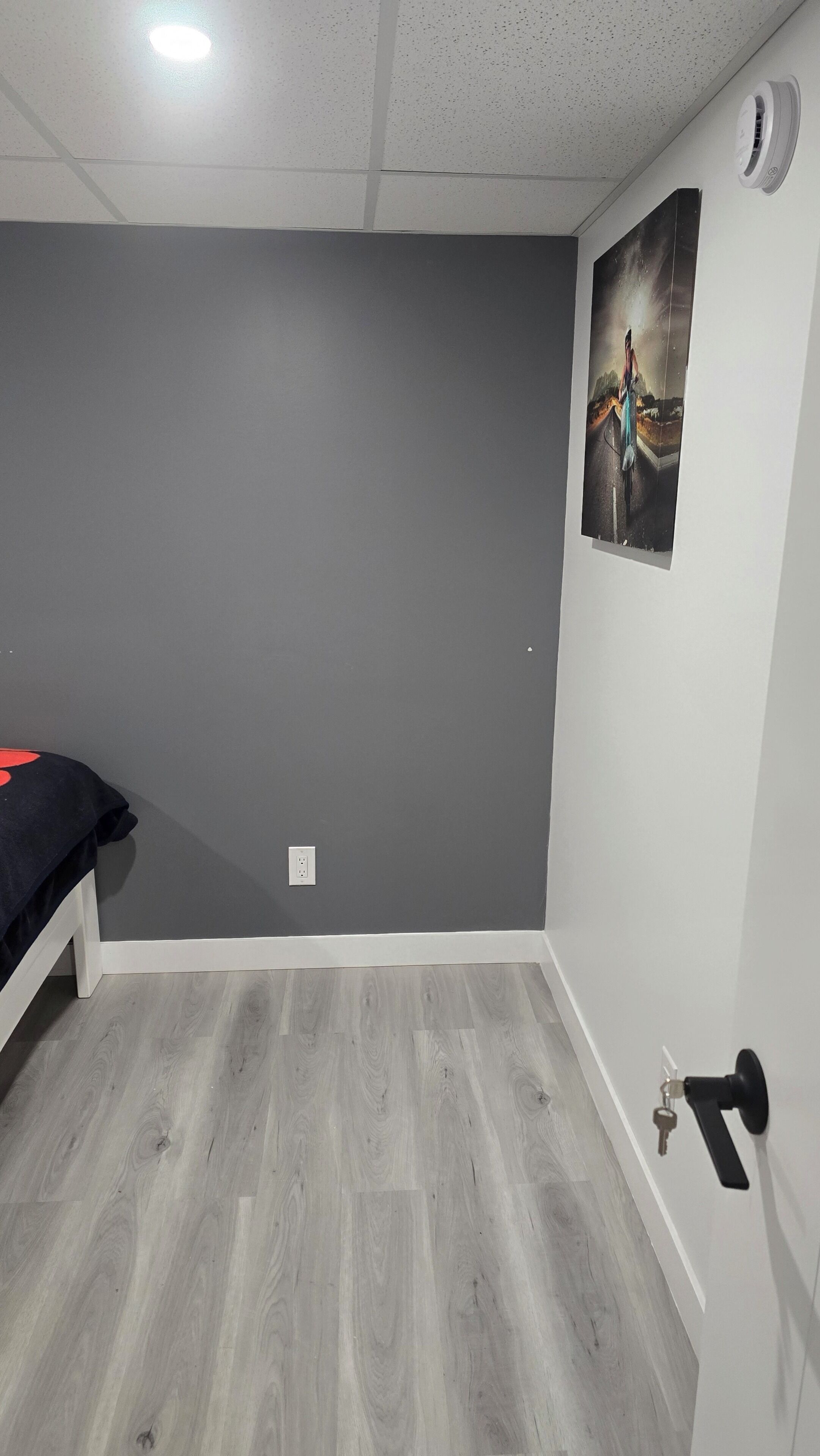 A room with grey walls, a wooden floor, a bed with a black cover, a door with a keyhole, and a wall-mounted air vent.