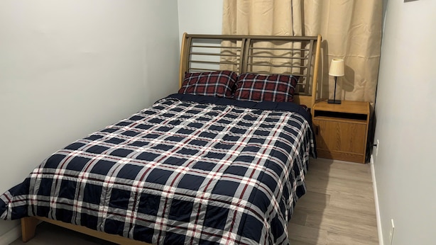 A single bed with a plaid bedspread, a wooden headboard, a nightstand with a lamp, and beige curtains.