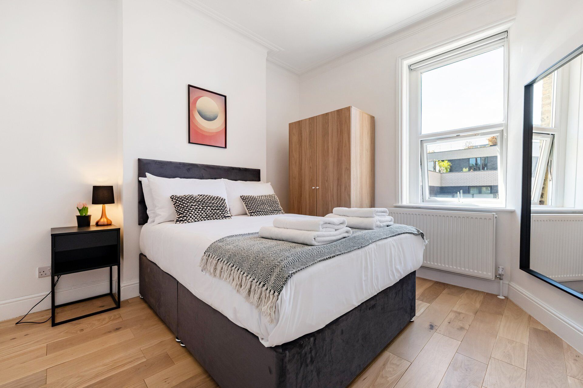 A modern bedroom with a bed, bedside table, lamp, artwork, wardrobe, and a window with a view of buildings.