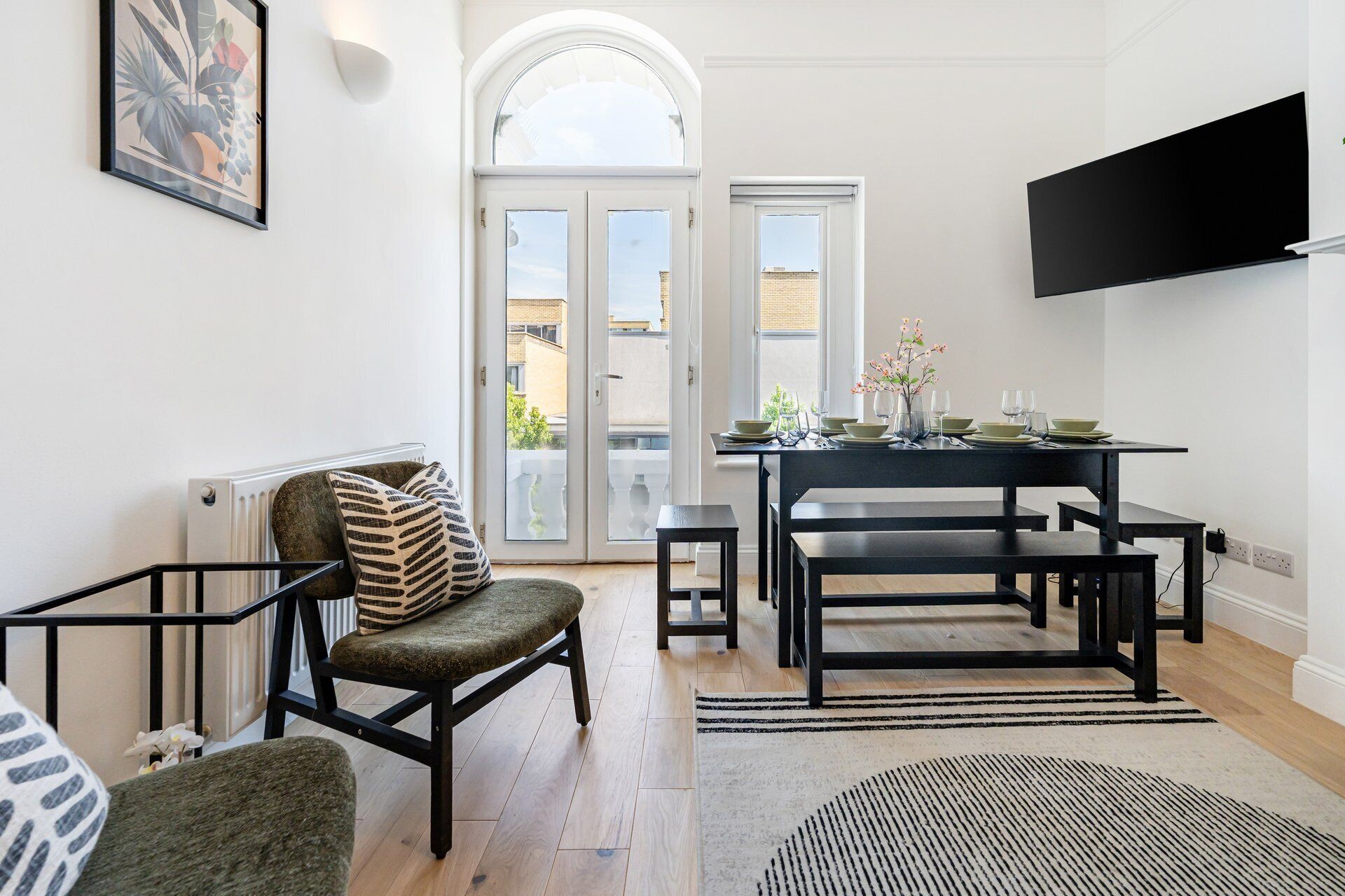A modern living room with a sofa, a dining table set for two, a television mounted on the wall, and a view through the door.