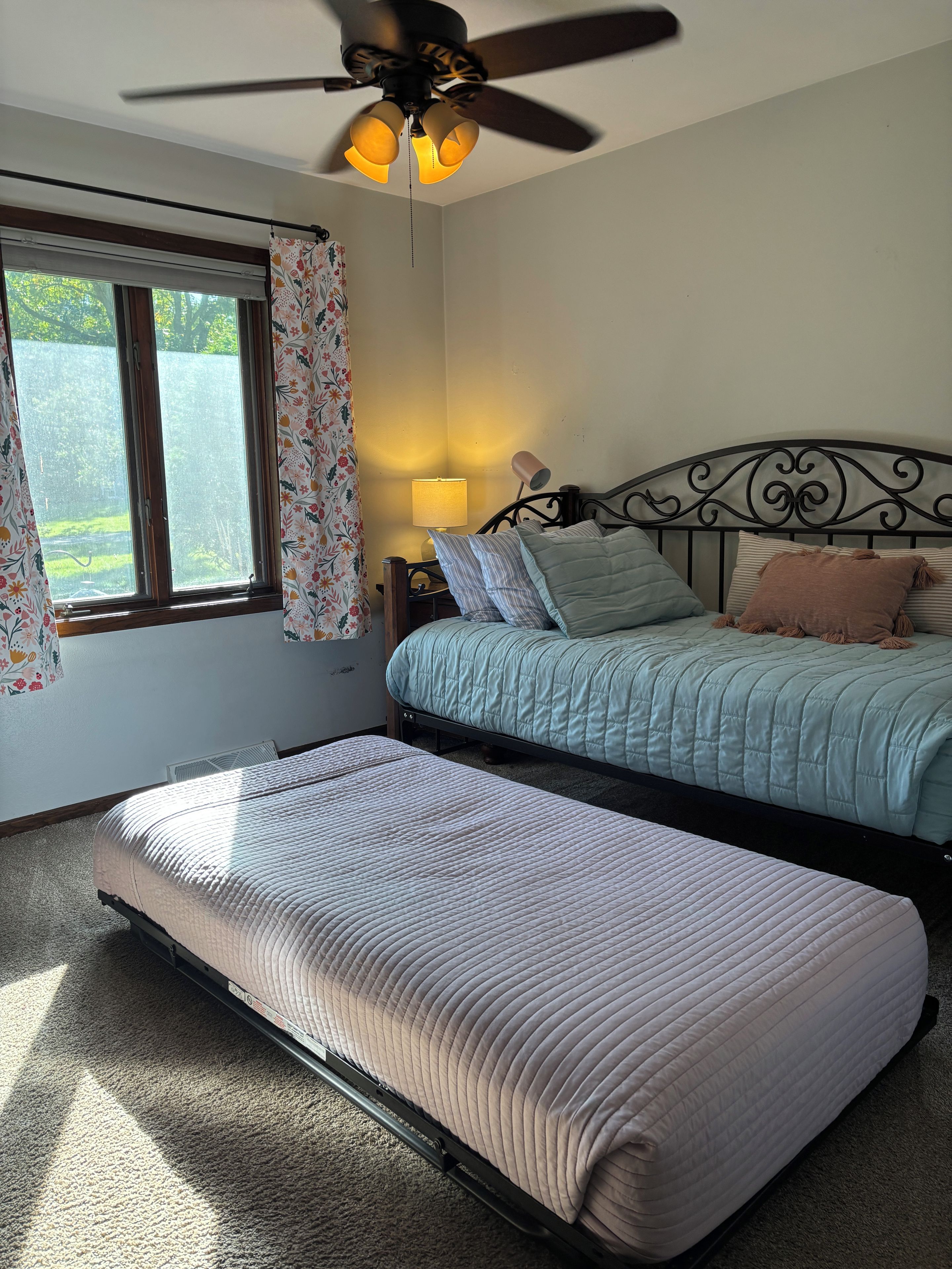 A bedroom with a bed, a bedside table with a lamp, a ceiling fan, and a window with curtains.