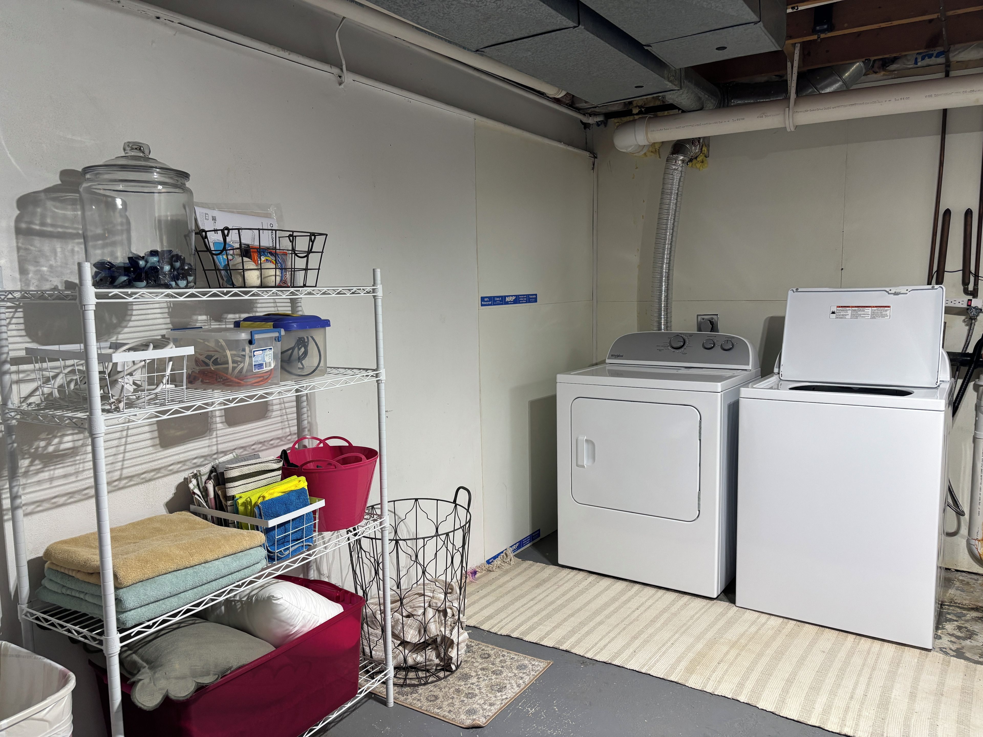 A laundry room with a washing machine, dryer, metal shelving with storage bins, and a utility room sign.