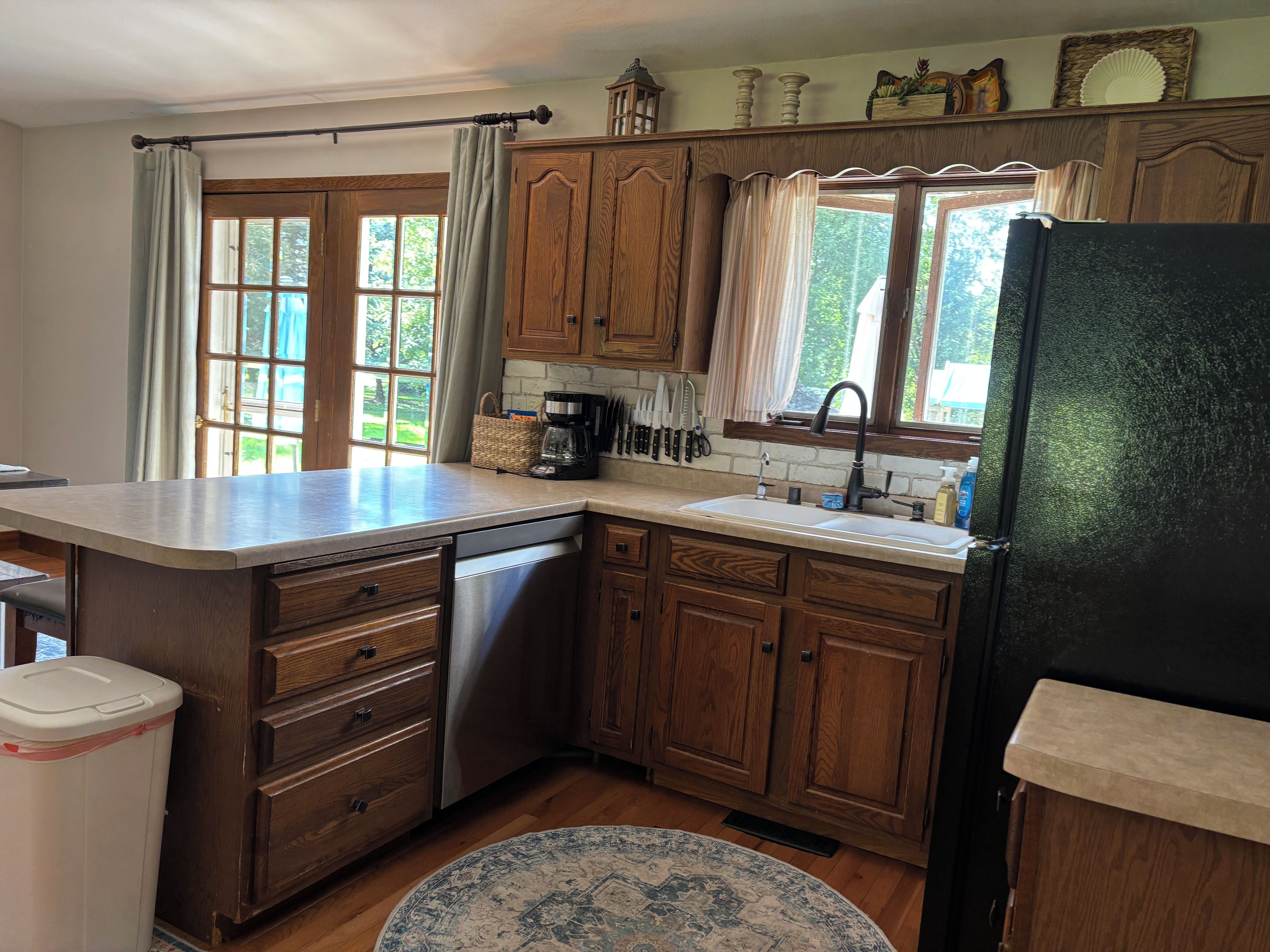 Una cocina con armarios de madera, lavavajillas de acero inoxidable, refrigerador negro y una ventana con cortinas.