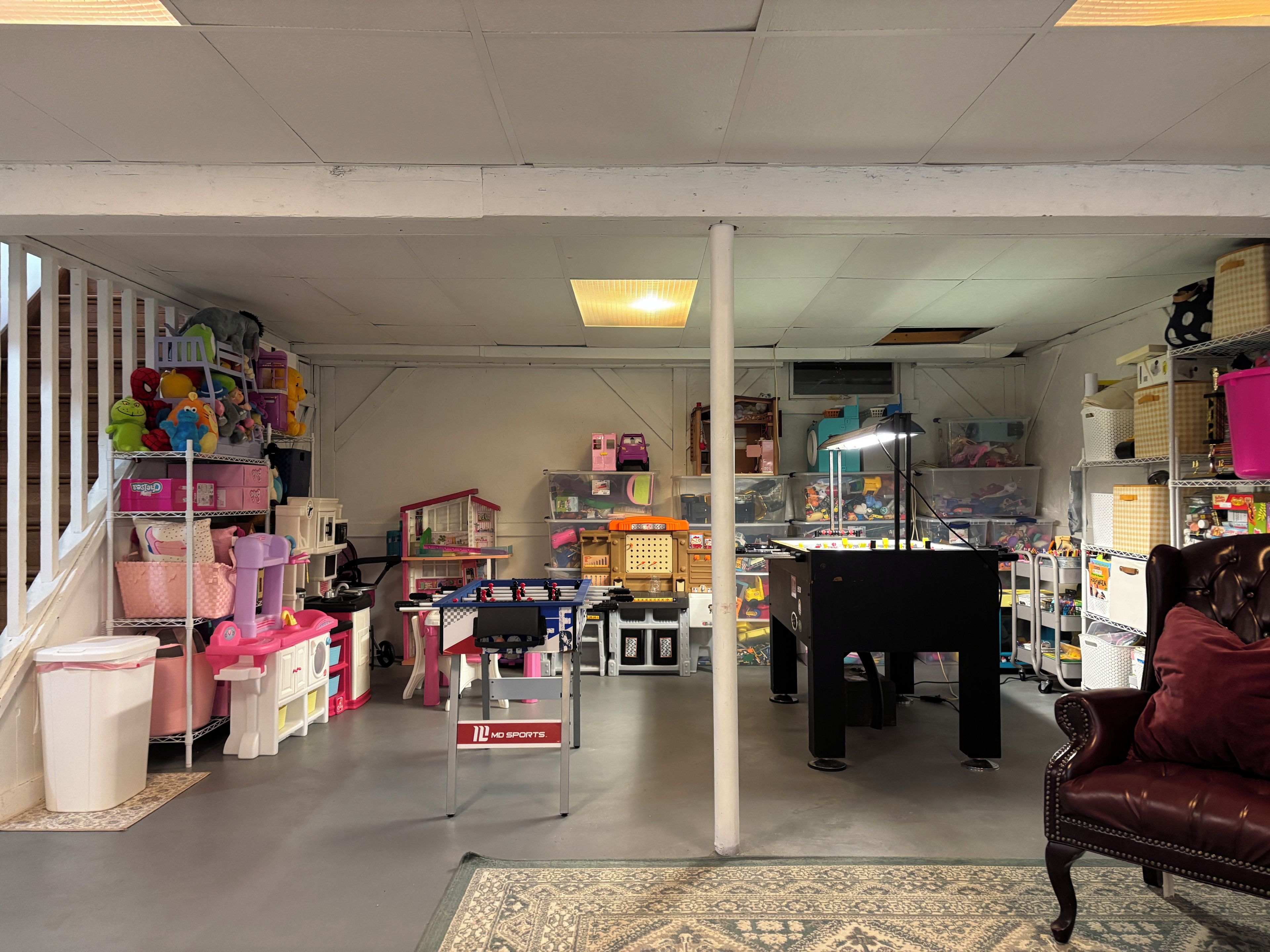 Et værelse med diverse legetøj, reoler med opbevaringskasser, et skrivebord med lampe og en læderstol.