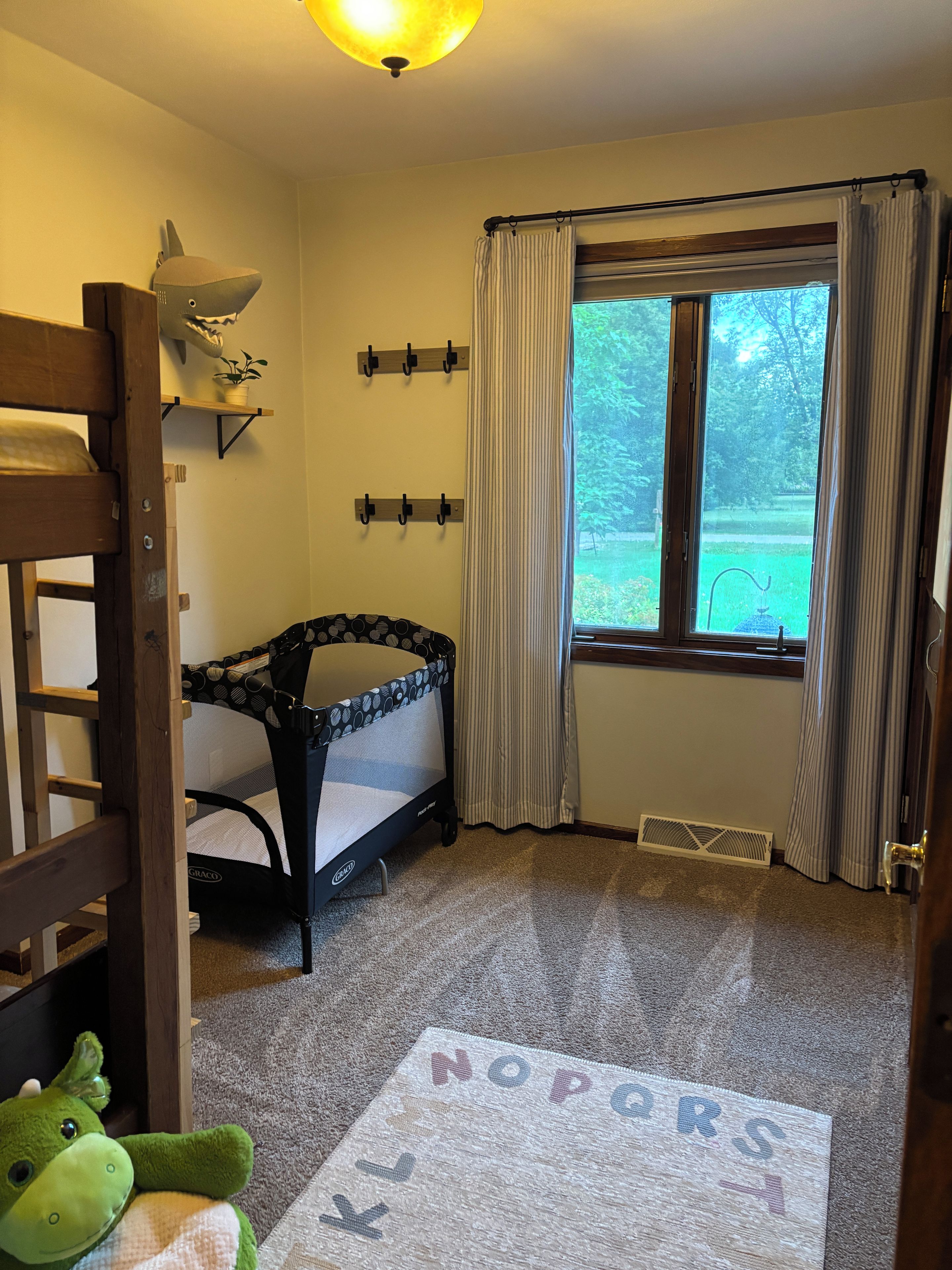 A child's room with bunk beds, a crib, a window with curtains, and a rug with a dinosaur design.