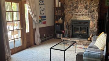 Amplia sala de estar con chimenea de piedra, ventilador de techo y un ventanal con cortinas.