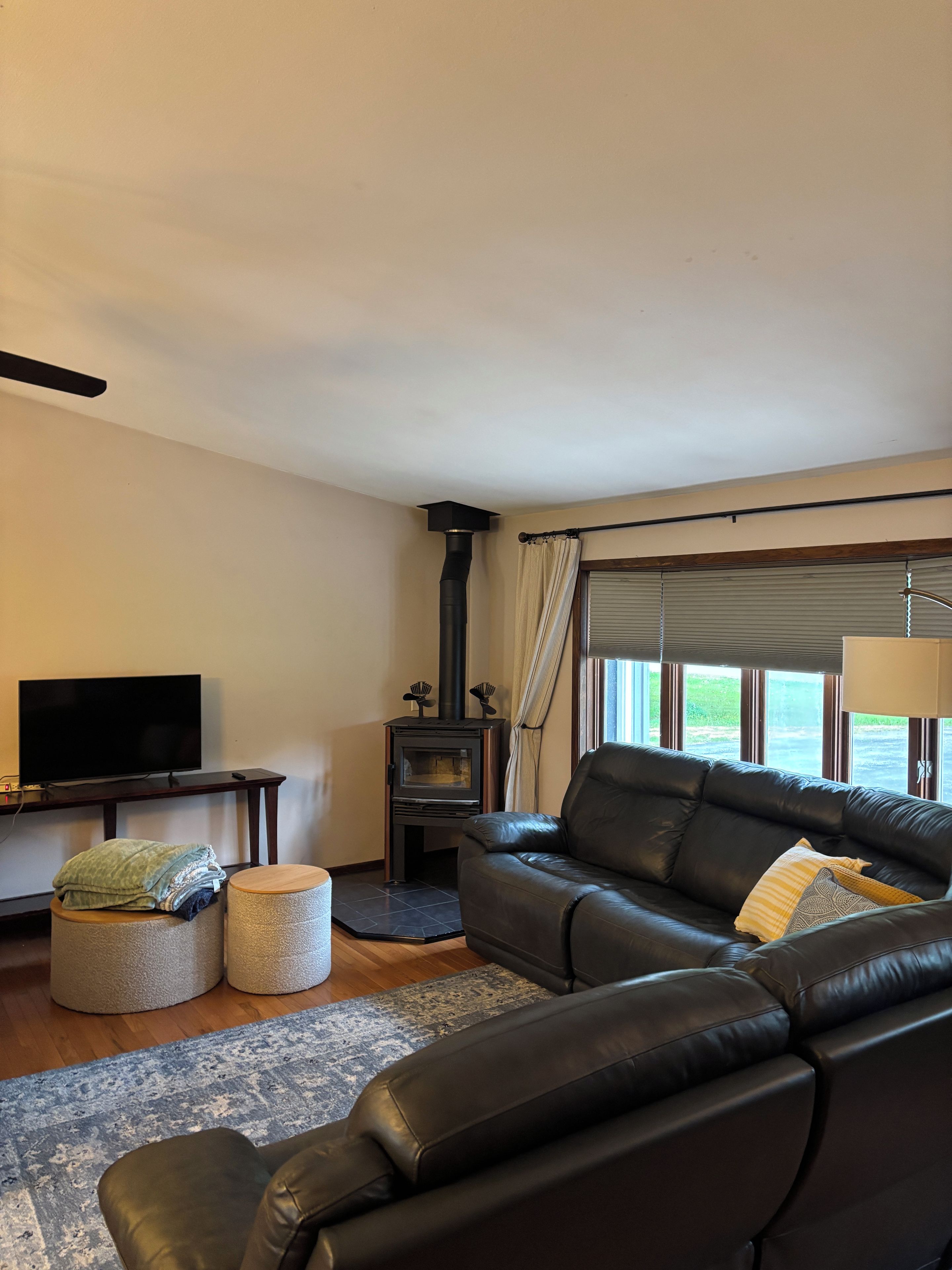 A living room with a black leather sofa, a flat-screen TV, a wood-burning stove, and a view through large windows.