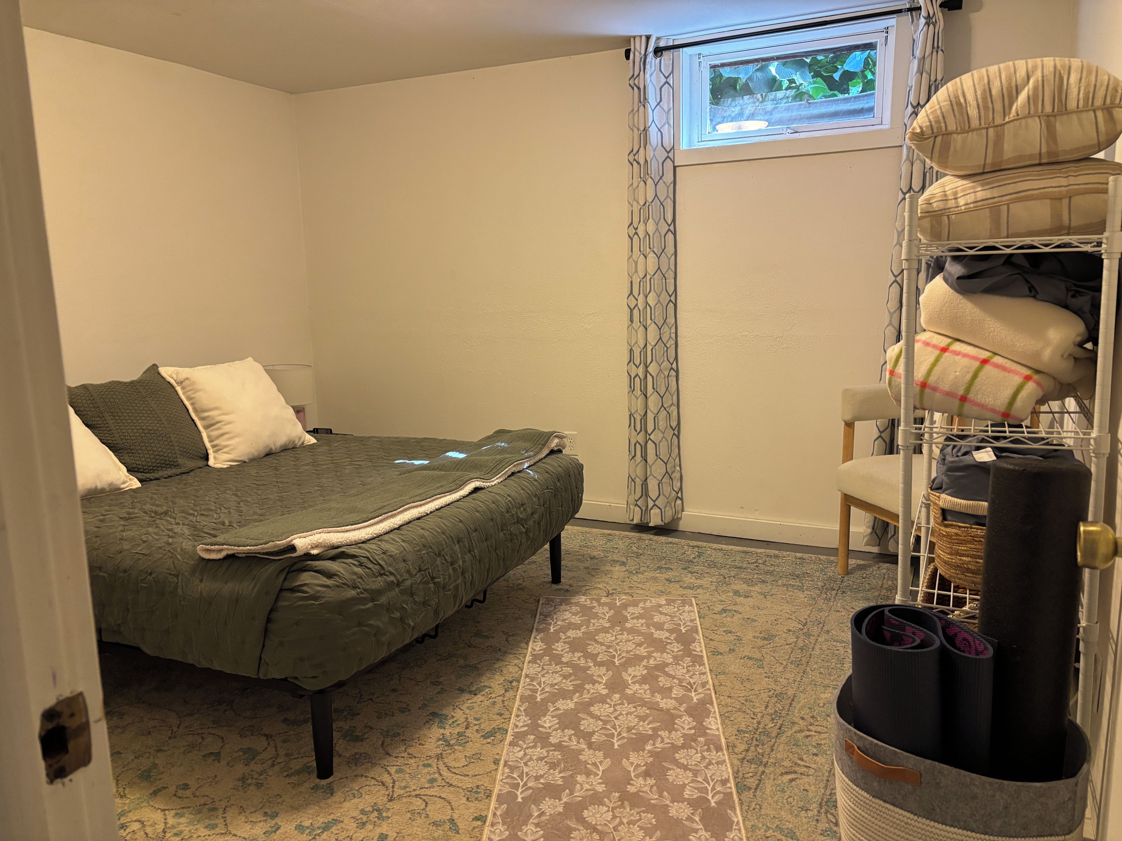 A bedroom with a bed, a chair, a shelf with folded blankets, and a window with a view of greenery.