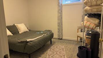 A bedroom with a bed, a chair, a shelf with folded blankets, and a window with a view of greenery.