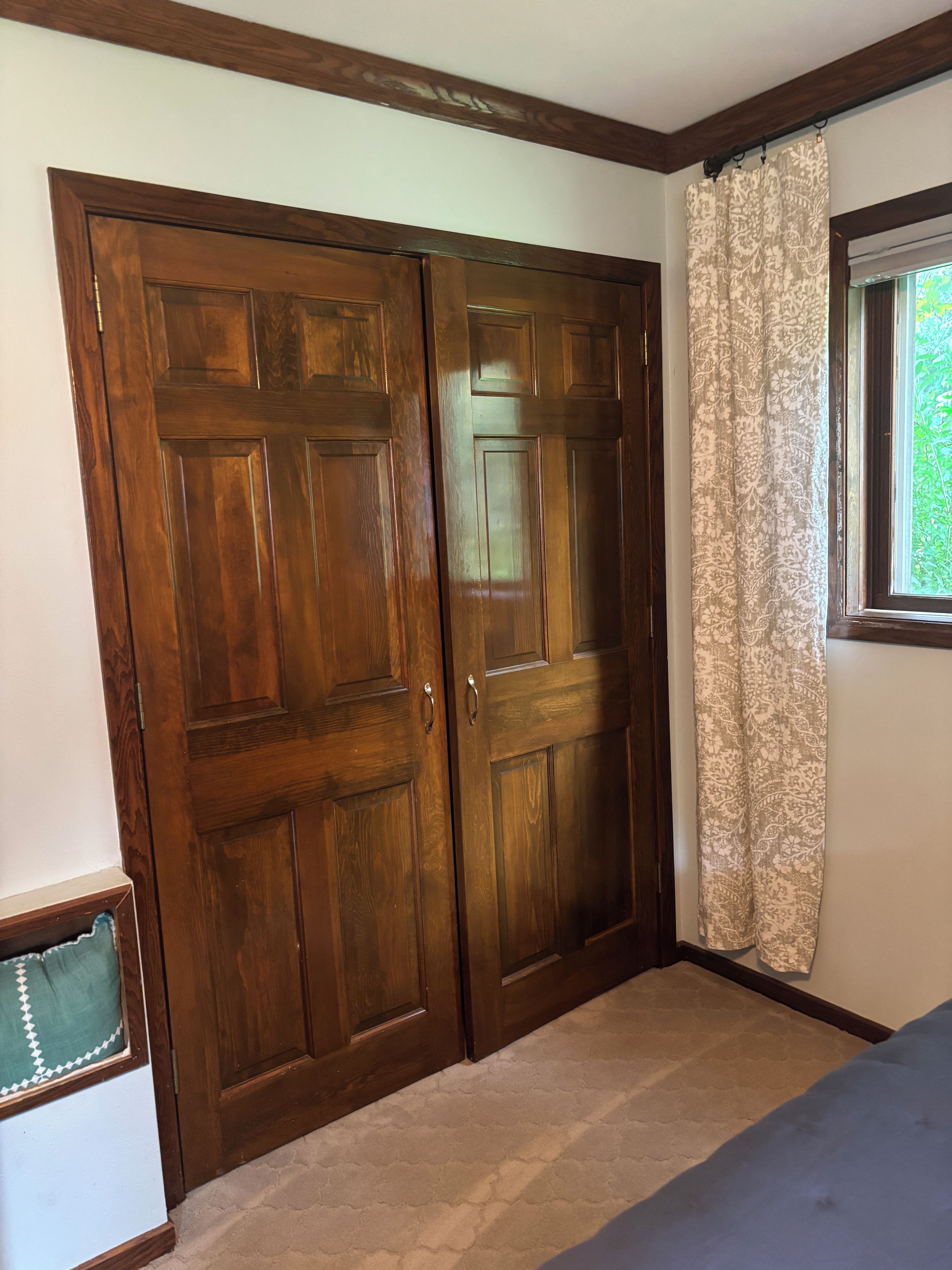 A room with a large wooden wardrobe, a window with patterned curtains, and a carpeted floor.