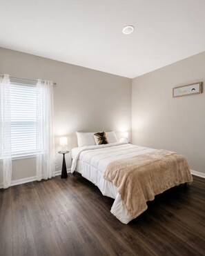 A bedroom with a bed, bedside table, lamp, and a window with curtains. - Bell Station Executive Cottage •Ground‑Level 1BR (Hurst)