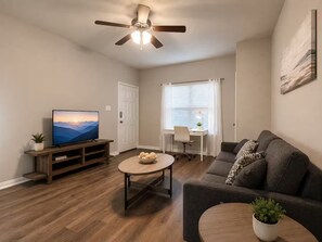 A living room with a television, a ceiling fan, a dining area with a table and chairs, and a sofa. - Bell Station Executive Cottage •Ground‑Level 1BR (Hurst)