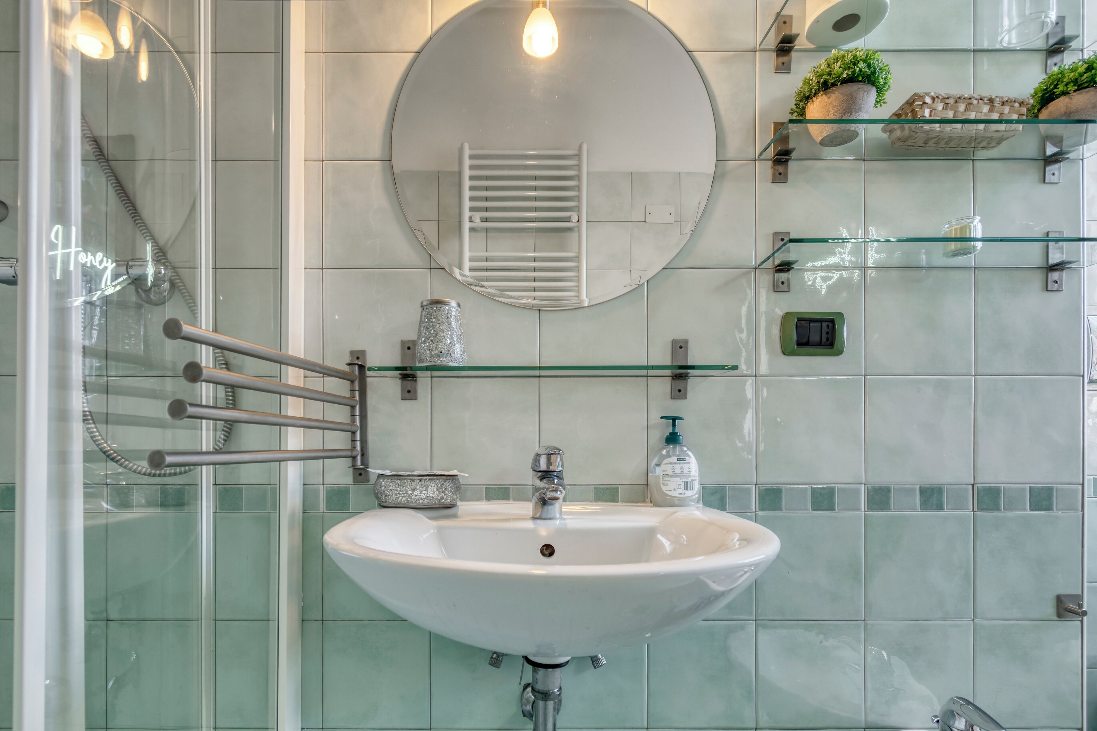 Een badkamer met een wit wastafel, een douchecabine van glas, een ronde spiegel en een glazen plank met toiletartikelen.