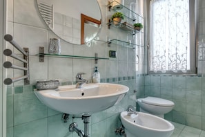 Un bagno con un lavandino bianco, un WC e un bidet. Sono presenti mensole in vetro con piante e uno specchio sopra il lavandino.