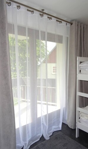 Quarto com janela coberta por cortinas finas, uma cama beliche branca e um tapete cinza no chão.
