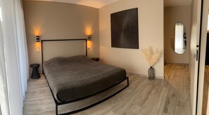 Een slaapkamer met een bed, nachtlampjes, een wanddecoratie, een spiegel en een vaas met gedroogde planten.