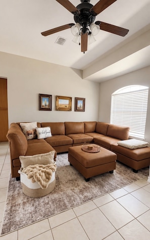 A living room with a brown sectional sofa, a round ottoman, a ceiling fan, and three framed pictures on the wall. - 4BR/2BA – Flexible Queen & Twin Sleeping – Quiet Neighborhood (Abilene)