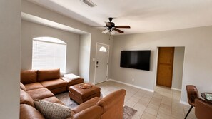 A living room with a sectional sofa, ottoman, and a flat-screen TV mounted on the wall. - 4BR/2BA – Flexible Queen & Twin Sleeping – Quiet Neighborhood (Abilene)