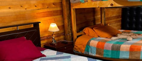 A cozy cabin room with a bunk bed, a single bed with a checkered quilt, a nightstand with a lamp, and wooden walls.