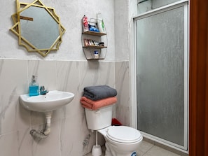 A bathroom with a white sink, a toilet, a shower with a glass door, a mirror with a geometric frame, and a shelf with toiletries.