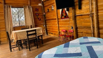 A cozy cabin room with a wooden interior, a bed with a blue and white quilt, a dining table with chairs, and a television mounted on the wall.