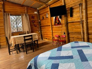A cozy cabin room with a wooden interior, a bed with a blue and white quilt, a dining table with chairs, and a television mounted on the wall.