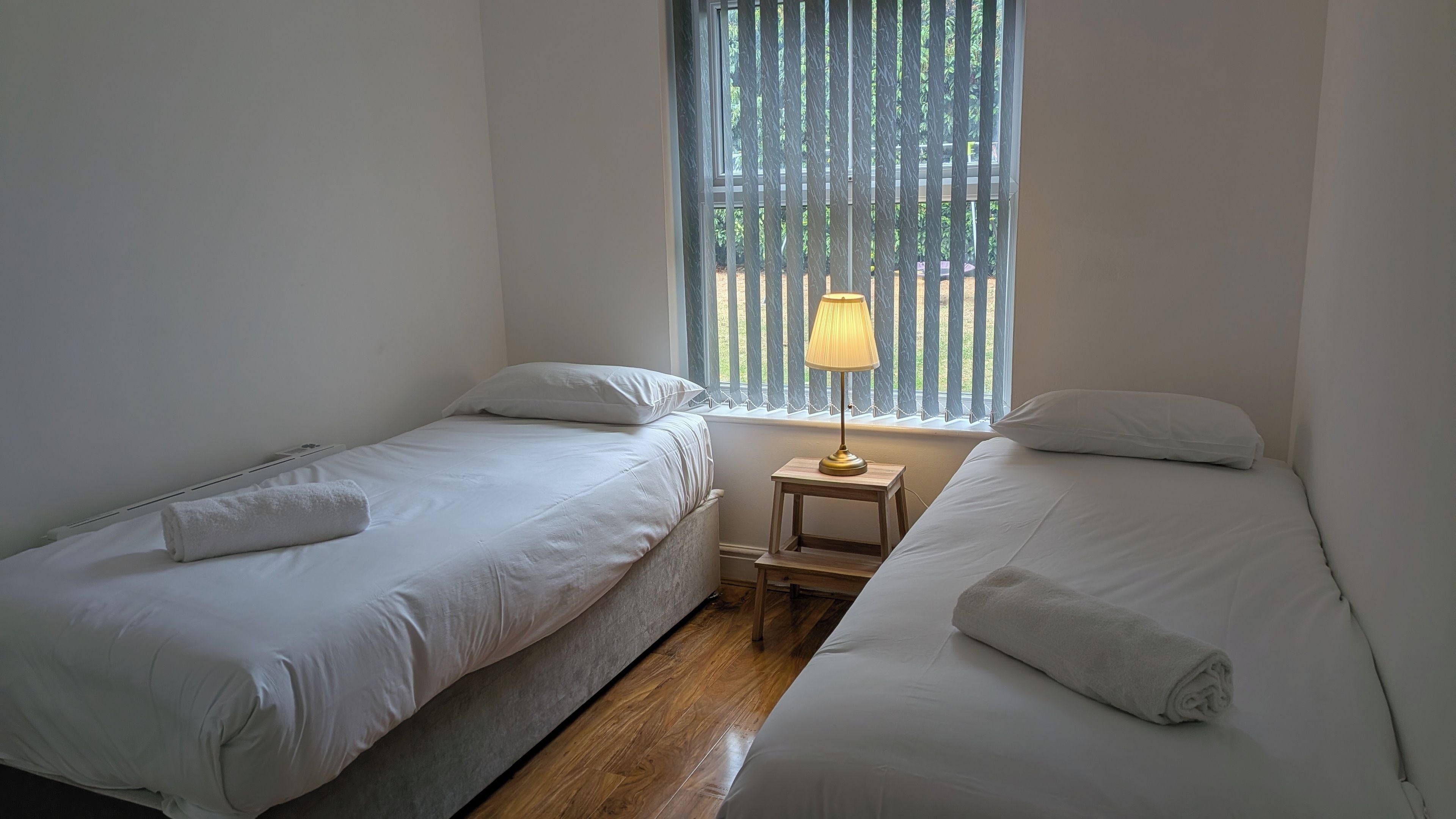 Una habitación con dos camas individuales, una mesita de noche pequeña con lámpara y una ventana con persianas verticales.