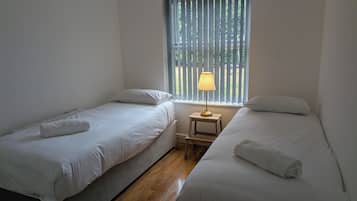 Una habitación con dos camas individuales, una mesita de noche pequeña con lámpara y una ventana con persianas verticales.