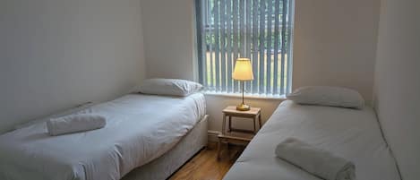 A room with two single beds, a small bedside table with a lamp, and a window with vertical blinds.