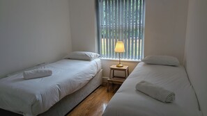 Una habitación con dos camas individuales, una mesita de noche pequeña con lámpara y una ventana con persianas verticales.
