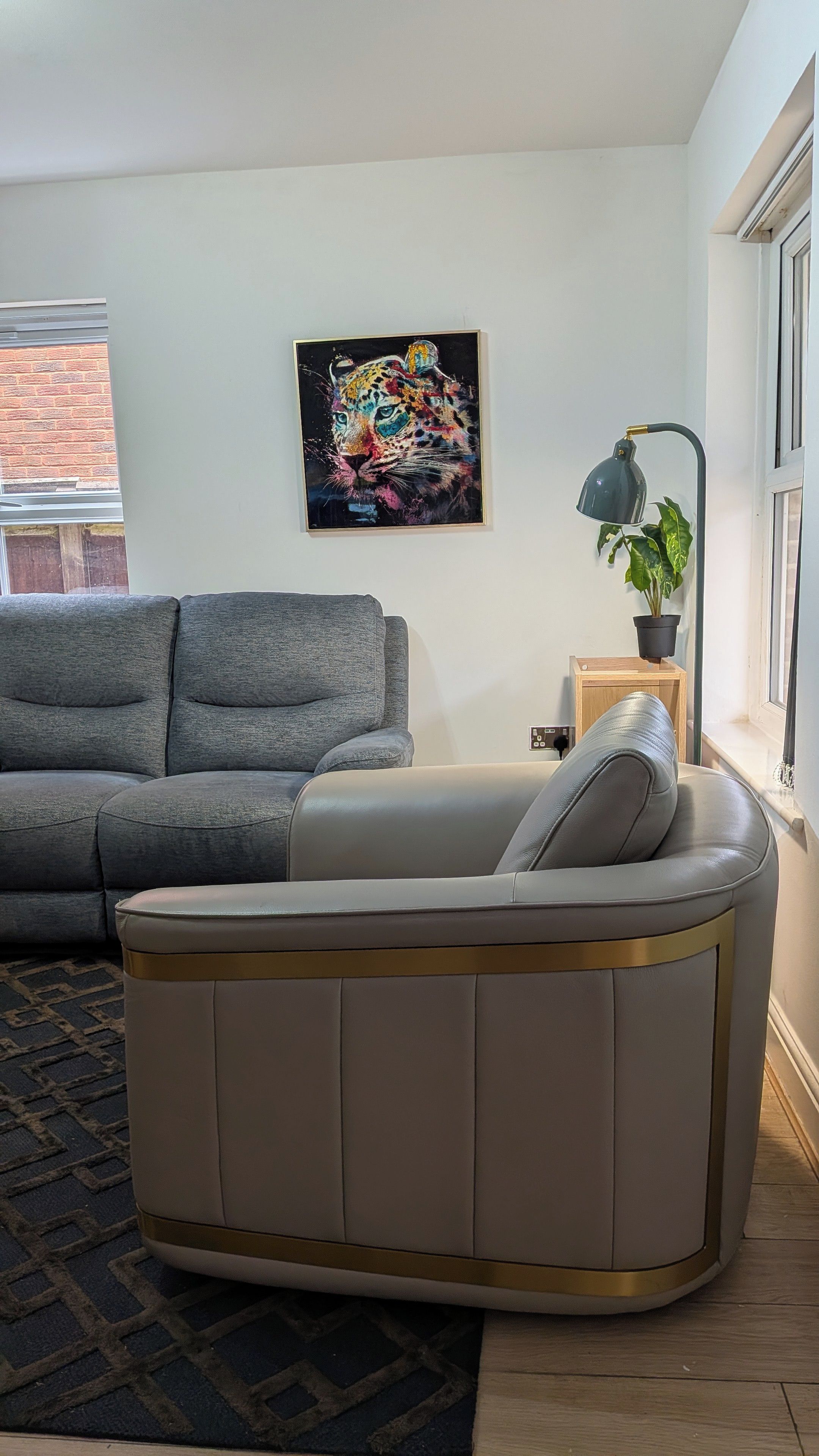 Een moderne woonkamer met een grijze bank, een hoekbank, een schilderij met zwarte lijst, een vloerlamp en een potplant.