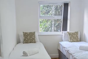 A room with two beds, each with a pillow and a rolled towel. There is a window with a view of trees.