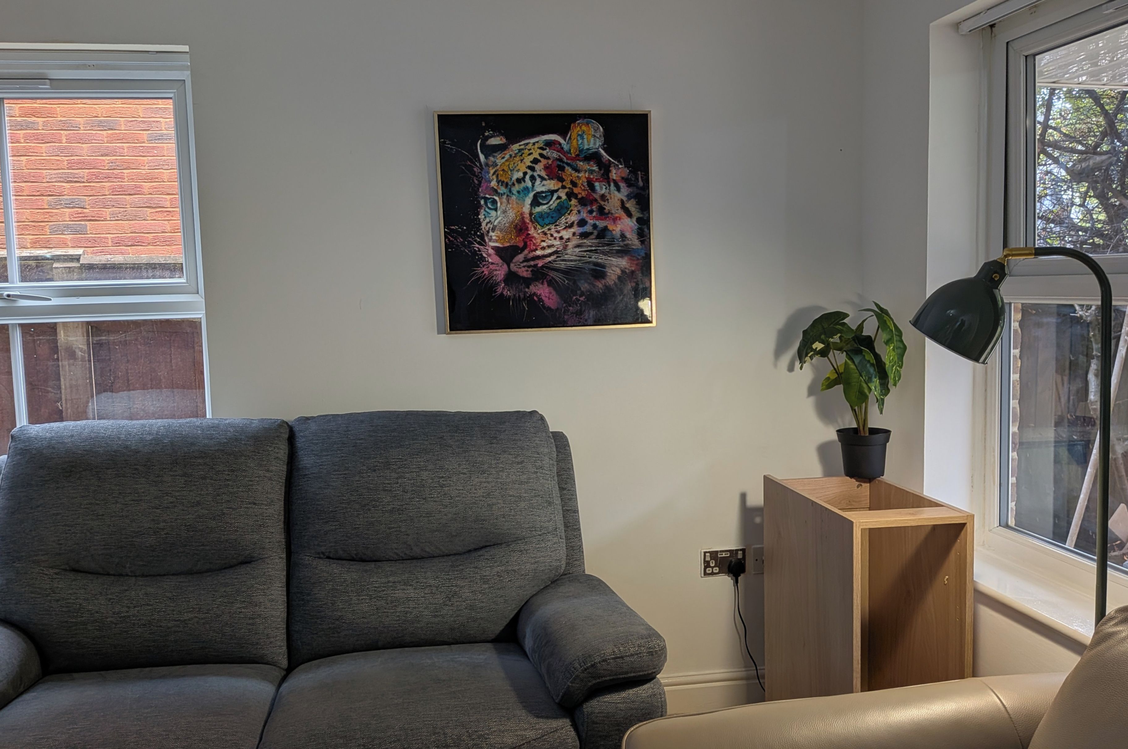 A modern living room with a grey sofa, a wooden side table with a plant, a black floor lamp, and a colorful abstract painting on the wall.