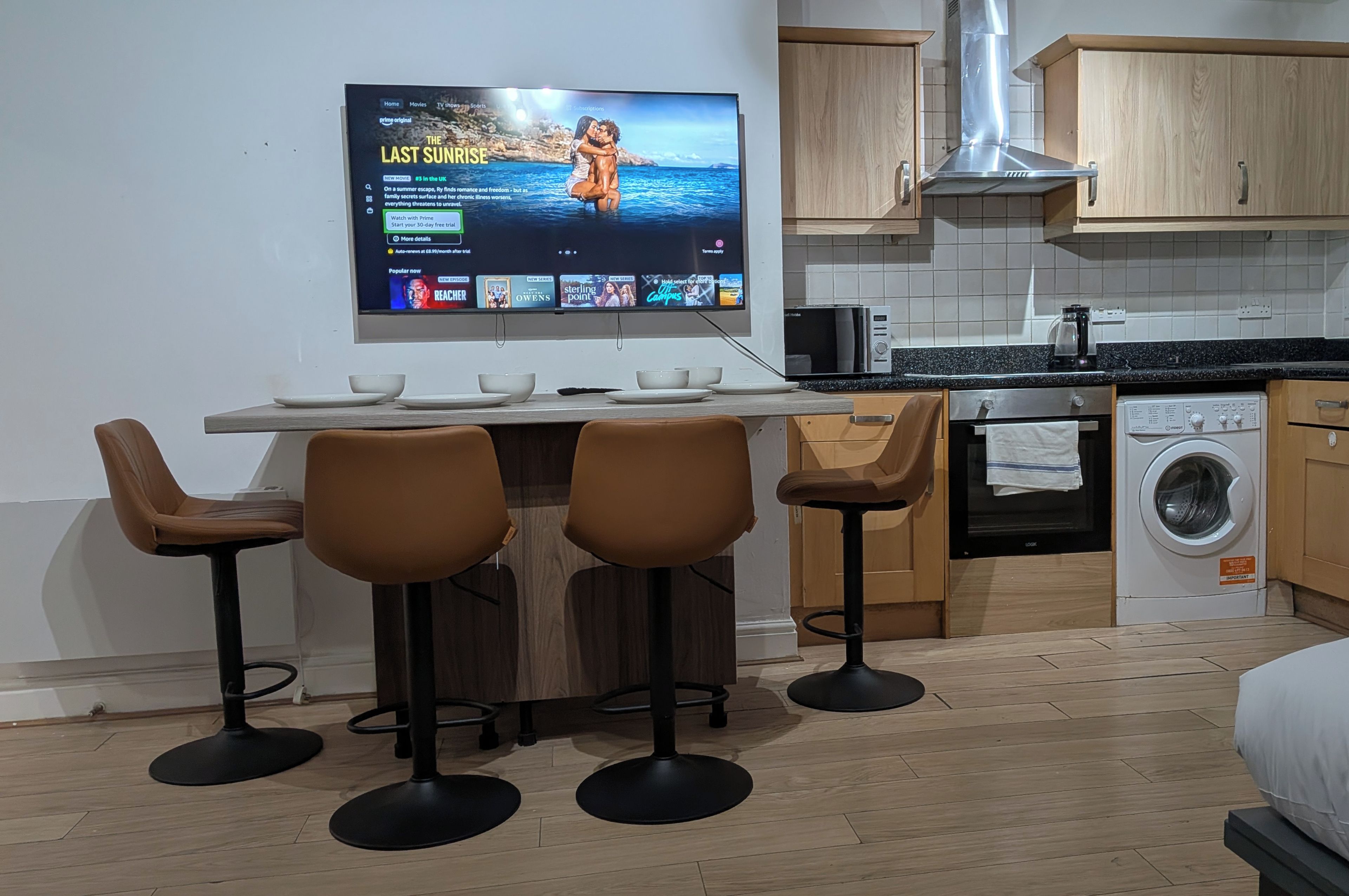 Une cuisine moderne dotée d’un téléviseur à écran plat fixé au mur, d’un coin repas avec une table et quatre tabourets de bar, d’un four à micro-ondes, d’une machine à café et d’un lave-linge.