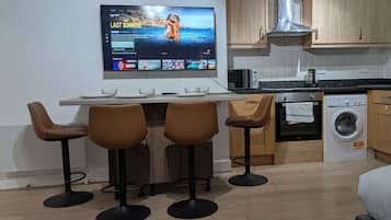 Une cuisine moderne dotée d’un téléviseur à écran plat fixé au mur, d’un coin repas avec une table et quatre tabourets de bar, d’un four à micro-ondes, d’une machine à café et d’un lave-linge.