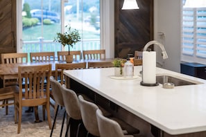 A modern kitchen with a large island, wooden chairs, a potted plant, and a view of a parking lot through the window. - Wings of Steel - 4BR Plus Bunk Room, Hot Tub, Fire Pit Table, 20 Minutes from Downtown Pittsburgh (Plum)
