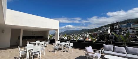 A rooftop terrace with outdoor seating, a kitchen area, and a view of a cityscape and mountains.