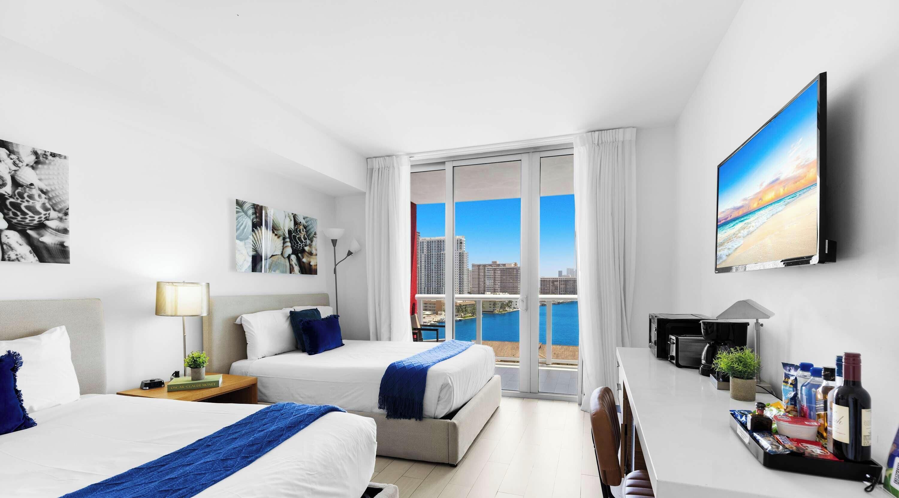 A hotel room with two beds, a desk, a TV, and a balcony with a city view.